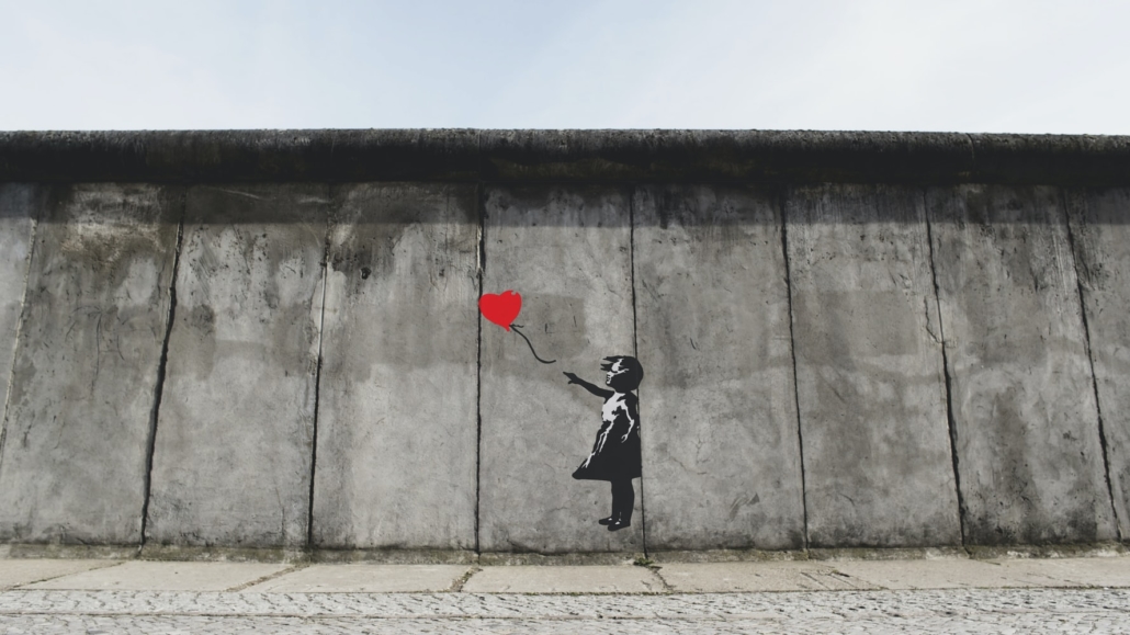 Pared de cemento con una niña en blanco y negro y un corazón rojo que parece que un globo y que la niña señala con el dedo.