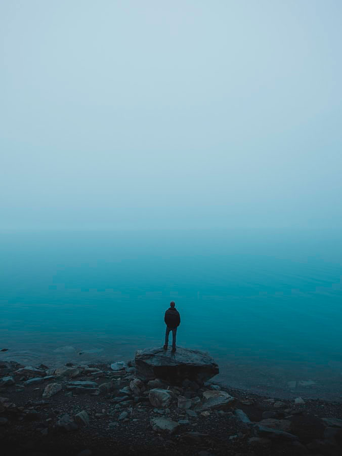 Hombre subido en una rato mirando hacia un lago azul. Sensación de tristeza y depresión. Blue day.